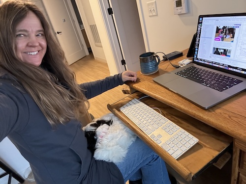 A girl with long hair sitting at a computer with a small fluffy white dog curled up on her lap