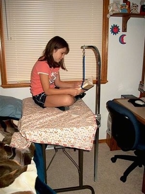 A girl is sitting on a grooming table in a bedroom and she is reading a book.