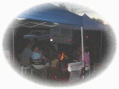 A family is sitting under an awning of a camper and there is a fire roaring in front of them.