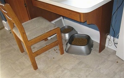 Two food Bowls under a kitchen table with a chair in front of them with scattered kibble on the floor