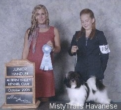 A girl is holding the leash of a dog standing on a table and in front of her. The dog is looking forward and to the left of them is a lady holding a blue ribbon.