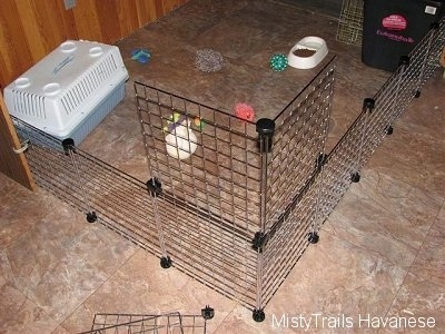 A wire rack enclosure that has a top rack being added to make it taller at the corner. There are dog toys, a food and water dish and a small carry type dog crate inside of the fenced off area.