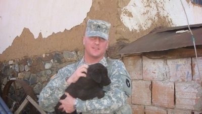 A Soldier dressed in camo is holding a black Sarplaninac puppy in his arms.