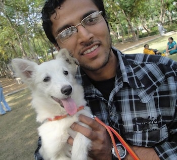 A white German Spitz puppy is being held in the air next to a man at a park. The German Spitz mouth is open and tongue is out