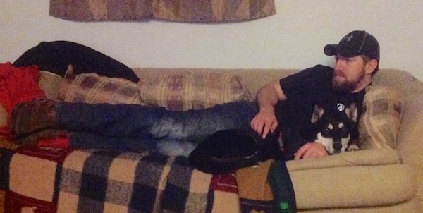 A black with tan and white Gerberian Shepsky is laying on a couch in front of a man in a black baseball cap, a black shirt, blue jeans and brown work boots, who has his arm around the dog.