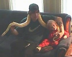 A man in a green Nike shirt is sitting next to a girl in a red sweat outfit. There is a Python around the mans neck and the tail is over the girl.