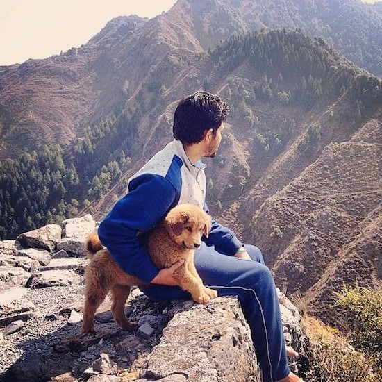 A man with dark black hair sitting outside at the edge of a cliff with a small tan puppy under his arm looking out at a good view of the mountains and valley.