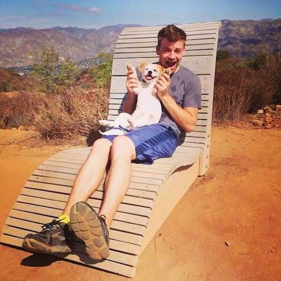 A man sitting on a wooden lawn chair outside in dirt with a view of the mountains behind them holding a small white with tan puppy belly-out smiling.