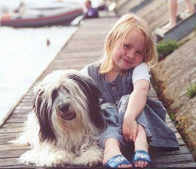 Front view - A grey and white with black Polish Lowland Sheepdog is laying on a wooden dock next to a girl sitting with blonde hair. They both are smiling. There is water next to them and a boat behind them.
