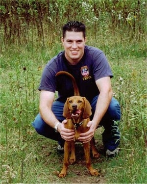 A man is kneeling behind a Redbone Coonhound puppy that is standing in grass. The puppy has its mouth open and it is licking the side of its mouth.