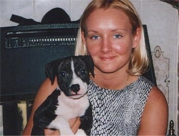 A blonde haired lady is holding a small black and white Staffordshire Bull Terrier puppy in her arms.