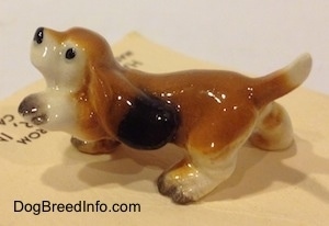 The left side of a tan with black and white ceramic Basset Hound figurine that has its front left paw in the air. The figurine has a short tail.