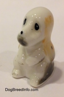 The front left side of a white with tan and black bone china Basset Hound puppy figurine that is sitting on its hind legs. The figurine has black circles for eyes.