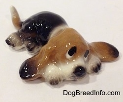 The front right side of a black and tan with white ceramic Basset Hound figurine that is laying down. The figurine has sad eyes.