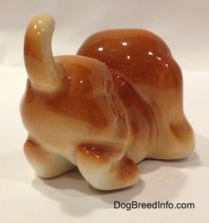 The back right side of a tan Cartoon style Bloodhound puppy. The figurine has a short arched up tail.