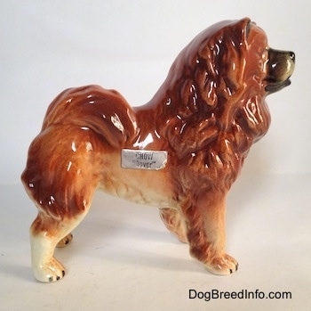 The right side of a brown with tan and black Chow Chow figurine. The figurine has a sticker across its side. The words - Chow 'lover' - is on the sticker. The dog has very small perk ears. Its tail is fluffy and curls up over its back.