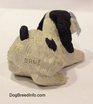 The back right side of a sandicast black and white American Cocker Spaniel that is sniffing a flower out of a knocked over pot figurine. The figurine has the letters - BRUE - on its back side.
