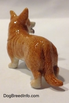 The back left side of a porcelain brown with white Cardigan Welsh Corgi figurine in a standing pose. The figurine has a large tail.