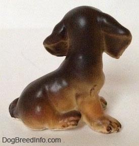 The right side of a brown with tan Dachshund puppy in a sitting pose figurine. The figurine has large brown ears.