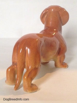 The back right side of a brown Dachshund figurine that is in a standing pose. The figurine has a long tail.