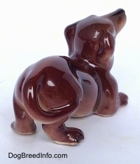 The back right side of a brown with tan Dachshund figurine that is in a play bow pose. The figurine is glossy.