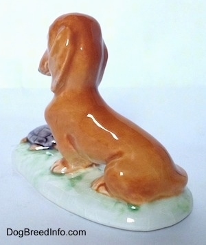 The back left side of figurine of a brown Dachshund in a sitting pose and there is a tortoise in front of it. Under the Tortoise and Dachshund is a grass platform.