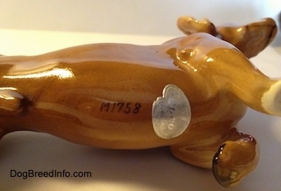 The underside of a porcelain Dachshund figurine. The figurine has a stamp that reads - M1758 - on its underside and to the right of that is a Napco Ceramic sticker.