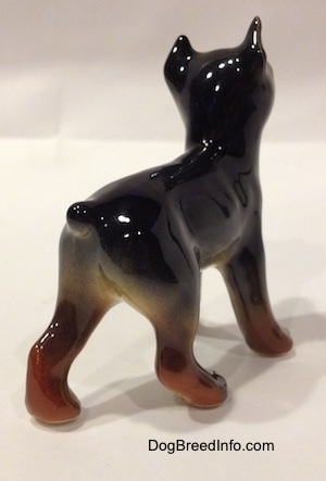 The back right side of a black and brown Doberman Pinscher puppy figurine. The figurine has a short tail.
