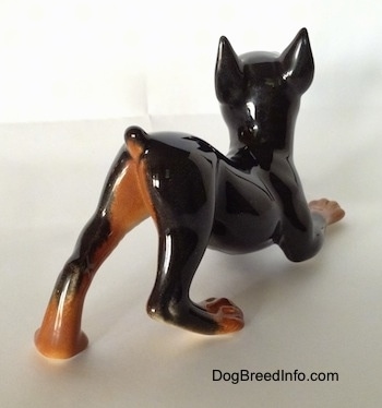 The back right side of a figurine that is of a Doberman Pinscher in a play bow pose. The figurine is glossy.