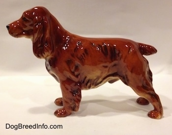 The left side of a red English Cocker Spaniel figurine. The figurines ears are long and hanging down to the sides. The tail is short. The nose is black and the snout is long.