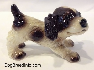 The right side of a liver and white English Springer Spaniel puppy figurine in a play bow pose. The tail of the figurine has a short liver colored tail and it is arched in the air.