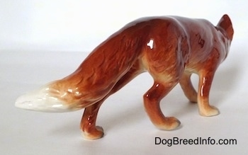 The back right side of a red fox figurine in a stalking pose. The figurine has a puffy long red tail, the end of the tail is white.
