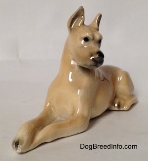 The front left side of a figurine of a tan Great Dane that is laying down. The figurine has its paws over top of each other and its ears are in the air.