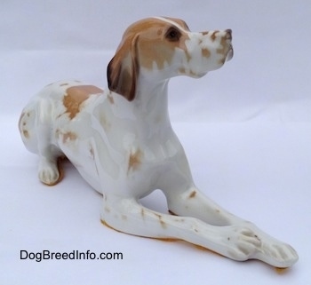The front right side of a porcelain white with brown figurine of a Pointer in a lying pose. The figurine has brown spots around its muzzle.