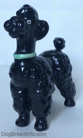 The front left side of a black Poodle with a green collar figurine. The figurine has a large white circle with a black dot for its eyes.