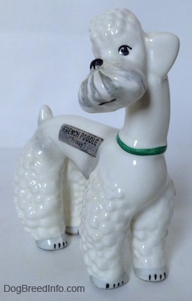 The front right side of a figurine of a white Poodle with a green collar on. The figurine has detailed black circles for eyes.