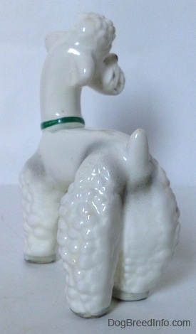 The back left side of a white Poodle figurine with a green collar on. The figurines leg has hair lumps on it.