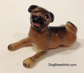 The front left side of a brown with black miniature Pug figurine lying down. The figurine has its mouth open and its head tilted to the right.