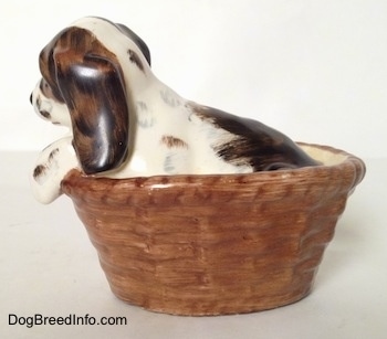 The left side of a figurine of a white with brown and black Russian Spaniel puppy in a basket. The figurine has a large brown with black spot above its tail.