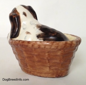 The back left side of a white with brown and black Russian Spaniel puppy in a basket figurine. The figurine has a brown with black spot on the back of its head.